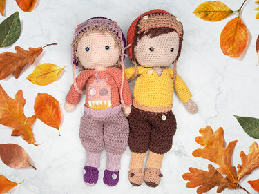 Zwei gehäkelte Amigurumi-Figuren mit Mützen und handgefertigter Kleidung, auf hellem Untergrund mit Herbstblättern gelegt.