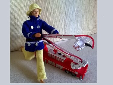 Puppe in blauer gestrickter Jacke, gelber Mütze und Hose neben rotem Spielzeug-Feuerwehrwagen