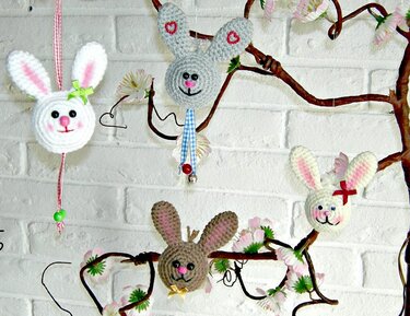 Mehrere gehäkelte Hasenköpfe in Weiß, Grau und Braun mit Schleifen und Verzierungen hängen an einem blühenden Zweig vor einer weißen Ziegelwand.
