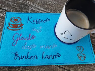 Türkisblauer Filz-Tassenteppich mit Bestickung ‚Kaffee ist Glück das man trinken kann‘ und Kaffeetassen-Motiven, darauf eine Tasse mit Katzenmotiv.