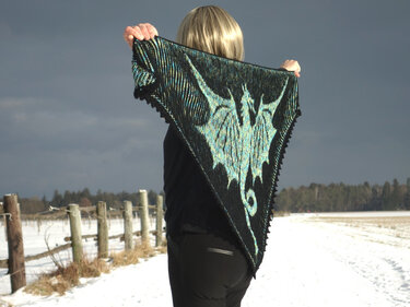 Person hält ein schwarzes, gestricktes Dreieckstuch mit türkisfarbener Drachen-Silhouette