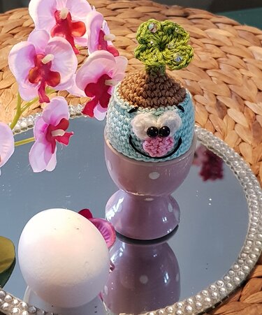 Hellblauer gehäkelter Eierwärmer mit Gesicht und kleinem grünen Baum auf lila gepunktetem Eierbecher, daneben ein weißes Ei und Orchideenblüten