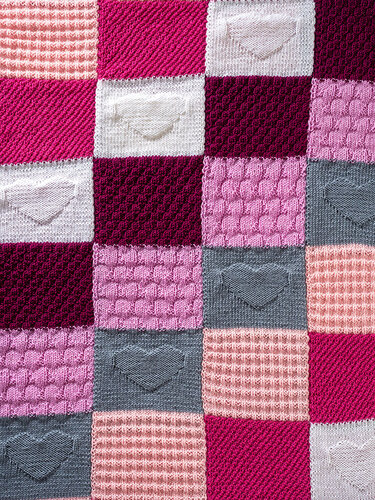 Nahaufnahme einer gestrickten Patchwork-Decke aus Quadraten in Rosa-, Bordeaux- und Grautönen mit Herz- und Noppenmustern