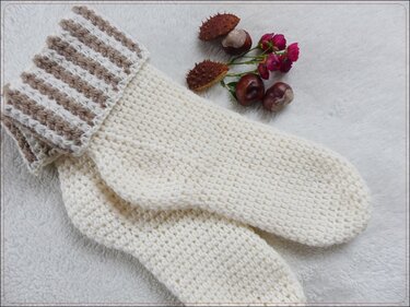 Zwei cremefarbene gehäkelte Socken mit braun-weiß gestreiftem Bündchen neben Kastanien und kleinen Blüten auf hellem Untergrund.