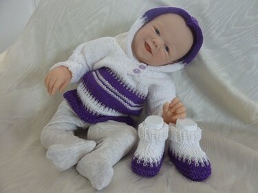 Babypuppe trägt gestricktes Ensemble mit Kapuze, Hose und Schühchen in Weiß und Lila