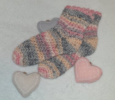 Handgehäkeltes Paar Socken in Rosa, Grau und Beige mit Wellenkante, liegend auf flauschigem weißen Untergrund neben drei Stoffherzen.