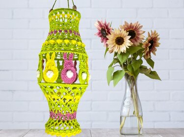 Gehäkelte grüne Laterne mit rosa und gelben Hasen-Applikationen und weißen Bommelschwänzchen neben einer Vase mit Sonnenblumen