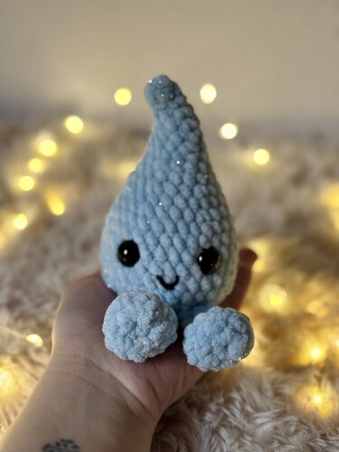 Hellblauer gehäkelter Amigurumi-Tropfen mit schwarzen Augen und gesticktem Lächeln, in einer Hand vor verschwommenen warmen Lichtern.