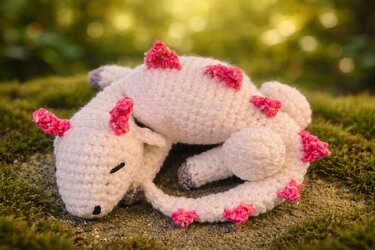 Gehäkelter weißer Amigurumi-Drache mit pinken Stacheln, zusammengerollt auf Moos