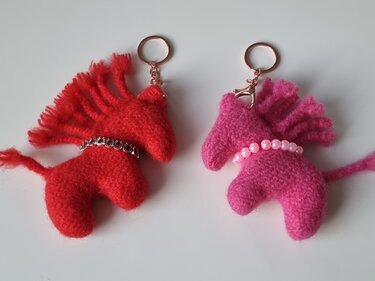 Zwei handgestrickte Pferde-Schlüsselanhänger, rot mit Hufeisenkette, pink mit Perlenkette, auf hellem Hintergrund.