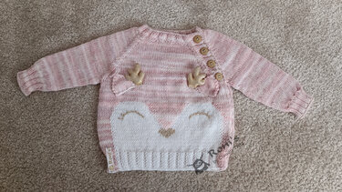 Rosa-weißer gestrickter Baby-Pullover mit Rehgesicht, goldfarbenen Applikationen und Schulterknöpfen, liegend auf Teppich