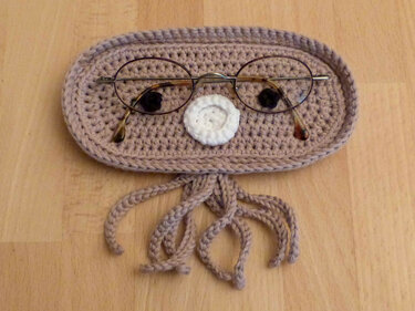Kompaktes, ovales Häkelutensilo in Beige mit weißer Applikation und Quasten, Brille liegt darauf.