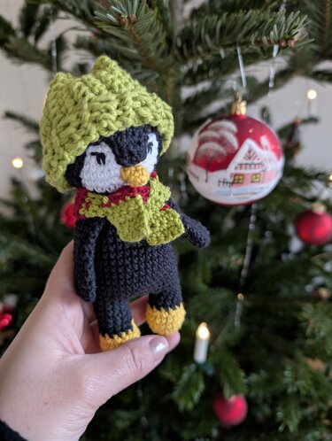 Gehäkelter Amigurumi-Pinguin mit grüner Mütze und Schal vor einem Weihnachtsbaum