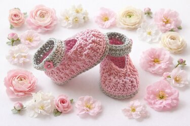 Rosa gehäkelte Babyschuhe mit graugrüner Umrandung und kleinem Knopf, arrangiert zwischen rosa und weißen Blüten auf weißem Hintergrund.