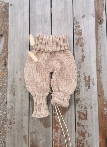 Beige handgestrickte Babyhose mit geripptem Bund und Bündchen auf hellgrauen Holzbrettern, dazu zwei getrocknete Ähren.