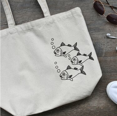 Naturfarbene Stofftasche mit schwarz gesticktem Motiv: drei stilisierte Fische und aufsteigende Blasen.