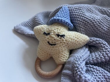 Gehäkelter gelber Stern mit aufgesticktem Gesicht, blauem Mützchen und rundem Holzring auf grauer Strickdecke