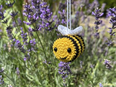 Nahaufnahme einer gehäkelten gelb‑schwarzen Biene mit weißen Flügeln, die an einem Faden vor verschwommenen lila Lavendelblüten hängt.