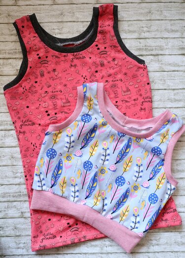 Zwei Kinder-Trägertops auf hellem Holzuntergrund: rosa Top mit „Little Princess“-Motiv und hellblaues Top mit Federn und Blumen