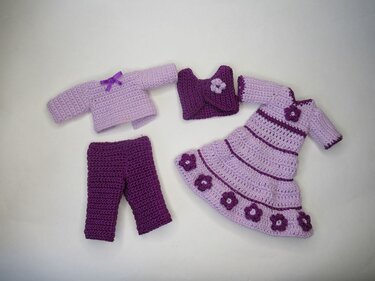 Vier gehäkelte Puppenkleidungsstücke in hellen und dunklen Lilatönen: Kleid mit Blüten, Hose, Bolero und Oberteil