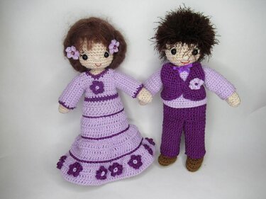 Zwei gehäkelte Amigurumi-Puppen in Flieder und Violett: langes Kleid mit Blumenapplikationen sowie Hose und Weste
