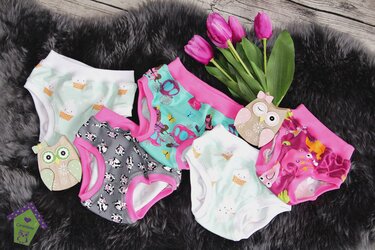 Bunte Baby- und Kinderunterhosen mit Eis-, Kuh-, Schmetterlings- und Meeresmotiven auf dunklem Kunstfell neben Holz-Eulen und pinken Tulpen.