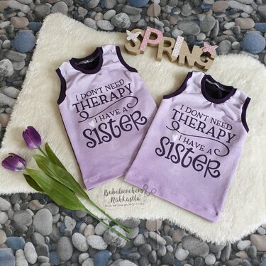 Zwei fliederne Kinder-Tanktops mit Schriftzug ‚I don't need therapy, I have a sister‘, dekoriert mit lila Tulpen und einem hölzernen SPRING-Schriftzug auf weichem Untergrund.
