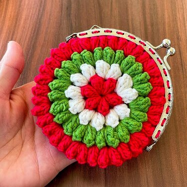 Hand holding round crocheted pouch with red, green and white floral motif and metal kiss-lock clasp.
