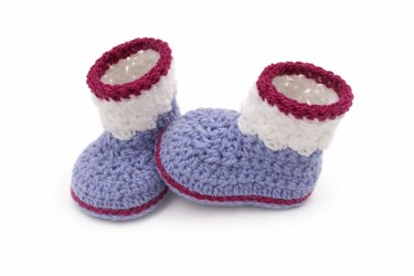Zwei handgehäkelte Babyschuhe in Lila, Weiß und Pink auf weißem Hintergrund.