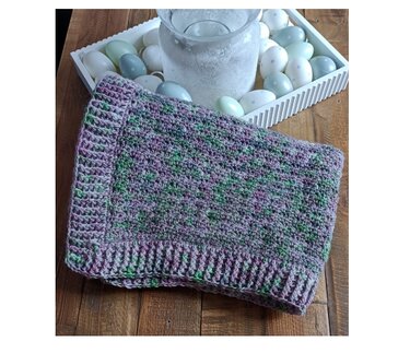 Gefaltete gehäkelte Babydecke mit geripptem Rand und violett-grünem Farbverlauf auf Holztisch