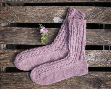 Paar fliederfarbene handgestrickte Zopfmustersocken auf rustikaler Holzfläche neben einer kleinen rosa Blüte.