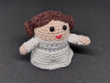 Gehäkelte Amigurumi-Figur mit braunen Haarknoten und weißem Kleid vor dunklem Hintergrund.