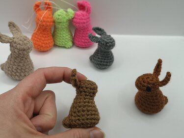 Mehrere mini gehäkelte Hasen in bunten Farben auf weißem Hintergrund, Hand hält einen braunen Hasen