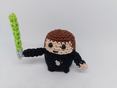 Gehäkelte Mini-Amigurumi-Figur mit braunen Haaren, schwarzem Körper und grünem Schwert auf weißem Hintergrund