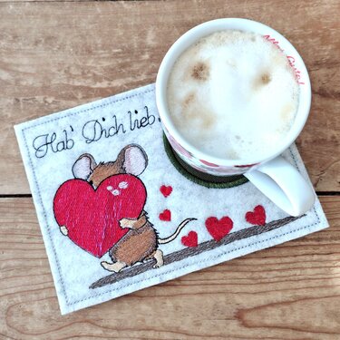 Bestickter weißer Tassenteppich mit einer braunen Maus, die ein rotes Herz hält, und dem Schriftzug "Hab' Dich lieb", teilweise von einer Tasse bedeckt.