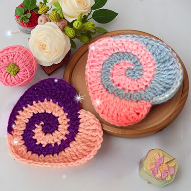Two spiral-pattern heart crochet pieces — one pink and gray, the other purple and peach — displayed on a wooden tray with cream roses and small crochet accents