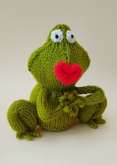 Green knitted frog figure with wide white eyes and a red heart-shaped mouth