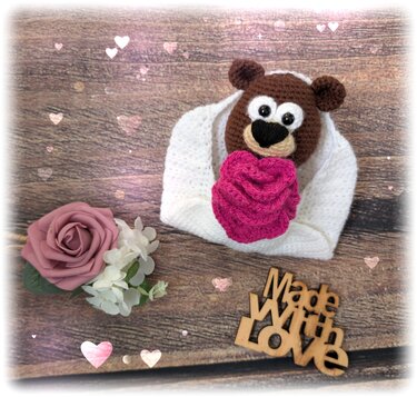 Gehäkelter brauner Amigurumi-Teddybär in weißer Decke hält pinkes Herz auf Holzuntergrund mit Blume