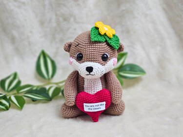 Niedlicher gehäkelter Otter mit gelber Blume auf dem Kopf hält rotes Herz