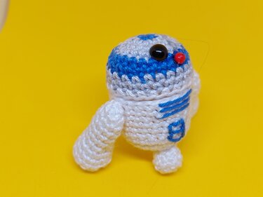 weiß-blaues gehäkeltes Amigurumi-Roboterchen vor gelbem Hintergrund