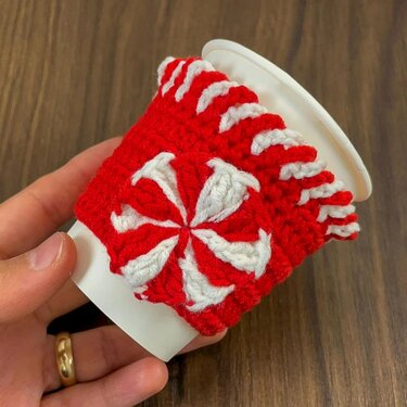 Red and white crocheted cup cozy with a peppermint swirl on a white paper cup held by a hand.