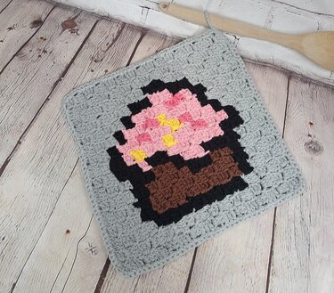 Square light grey pixel crochet potholder with a cupcake motif on a wooden surface