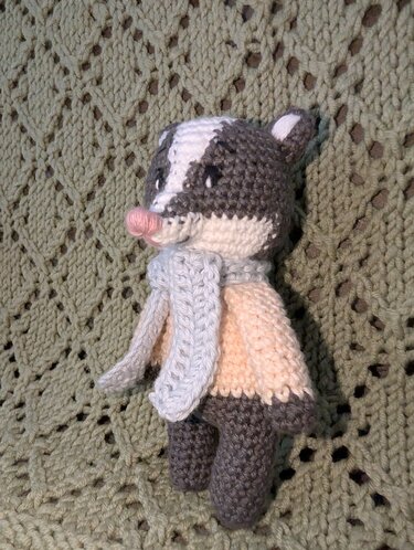 Gehäkeltes Dachs‑Amigurumi in Grau, Weiß und Creme mit hellblauem Schal auf gestrickter Decke