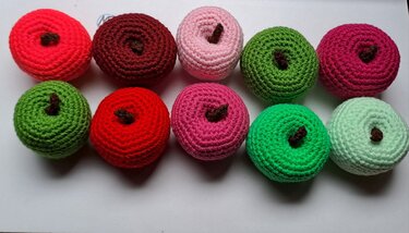 Zehn gehäkelte Äpfel in Rot-, Pink- und Grüntönen mit braunen Stielen auf weißem Hintergrund