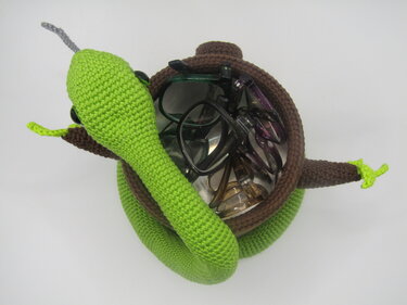 Gehäkelter brauner Korb in Schildkrötenform mit einer grünen Schlange, gefüllt mit mehreren Brillen.
