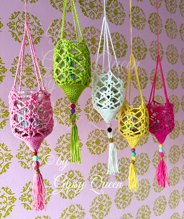 Fünf bunte gehäkelte Hüllen für Teelichtgläser mit Perlen und Quasten, hängend vor rosa Wand mit goldfarbenem Muster.