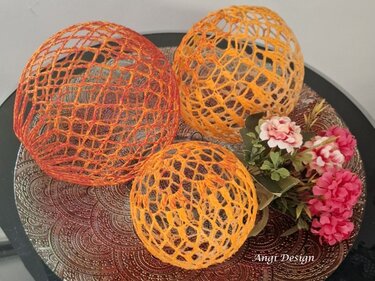 Drei gehäkelte offene Kugellampions in Gelb- und Orange-Tönen auf verzierter Platte neben Blumendekor