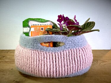 Gefilzter Häkelkorb in Rosa und Grau mit integrierten Griffen, gefüllt mit einer violetten Topfpflanze und verpacktem Gebäck auf einer Holzoberfläche.