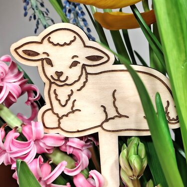 Holzstecker in Form eines liegenden Lamms mit Gravurlinien zwischen Frühlingsblumen