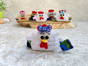 Mehrere gehäkelte Hühner mit verpackten Süßigkeiten auf einer Holzscheibe vor hellem Stoff